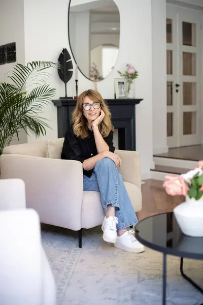 現代の家でアームチェアに座って魅力的なブロンドの髪の女性 黒のシャツとジーンズを着た笑顔の女性 ロイヤリティフリーのストック写真
