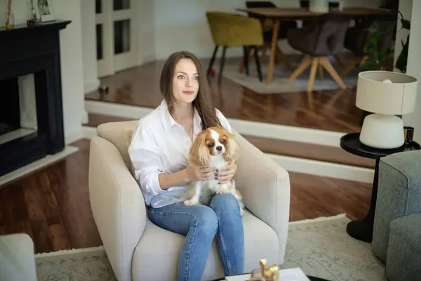 現代の家でアームチェアでリラックスする彼女のかわいい子犬と美しい長髪の女性の肖像画 アメリカ合衆国 ロイヤリティフリーのストック画像