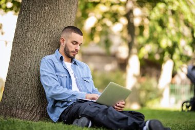 Genç adam dışarıda çalışmak ya da çalışmak için dizüstü bilgisayar kullanıyor. Genç bir erkek günlük kıyafetler giyiyor ve halka açık bir parkta çimlerin üzerinde oturuyor. Tam boy çekim.