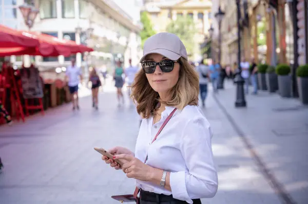 魅力的な女性が街の歩道やテキストメッセージを歩いています 白いシャツと黒いズボンを着ている中年女性 全長ショット — ストック写真