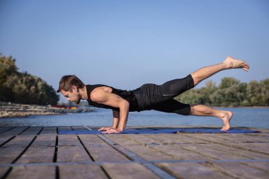 Orta yaşlı bir adam dışarıda yoga yapıyor. Kafkasyalı adam yoga minderi kullanıyor ve kalas pozisyonu tutuyor..