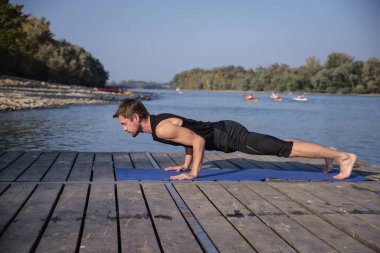 Orta yaşlı bir adam dışarıda yoga yapıyor. Kafkasyalı adam yoga minderi kullanıyor ve kalas pozisyonu tutuyor..