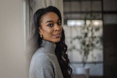 Pencerenin önünde durup kameraya bakan Afro-Amerikan bir kadının yakın plan portresi..