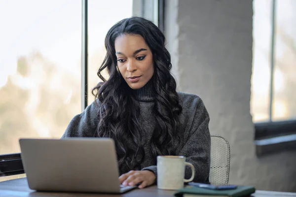 アフリカ系アメリカ人女性が窓際のテーブルに座ってノートパソコンに取り組んでいる ホームオフィス カジュアルな服を着ている魅力的な女性 — ストック写真