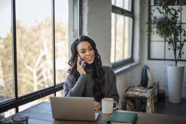 窓からテーブルに座ってスマートフォンで話すアフリカ系アメリカ人女性 ホームオフィス — ストック写真