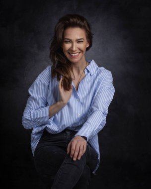 Çekici, orta yaşlı, dişlek, mavi tişört ve siyah kot pantolon giyen, izole, koyu arka planda oturan bir kadın. Uzayı kopyala Stüdyo çekimi.