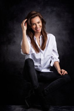 İzole edilmiş koyu gri arka planda oturan beyaz dişli, gülümseyen güzel bir kadın. Beyaz gömlek ve siyah kot pantolon giyen mutlu bir kadın. Boşluğu kopyala.