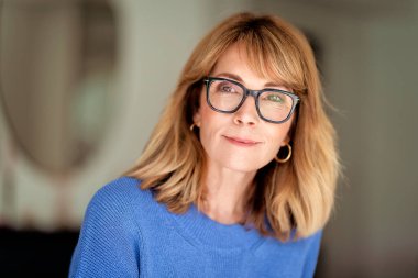 Kameraya bakıp gülümseyen orta yaşlı sarışın bir kadının portresi. Gözlüklü ve mavi gömlekli kendinden emin bir kadın.. 