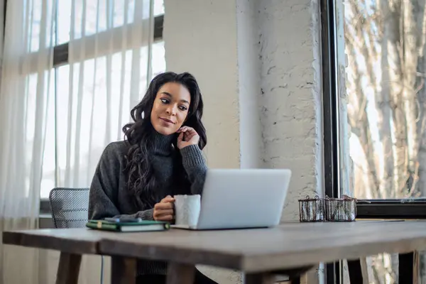 アフリカ系アメリカ人女性が窓際のテーブルに座ってノートパソコンに取り組んでいる ホームオフィス ウェブカンファレンスを開催する魅力的な女性 ロイヤリティフリーのストック画像