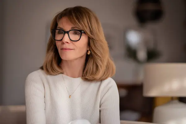 自宅のアームチェアでリラックスする魅力的な中年女性のクローズアップ ブロンド髪の女性は眼鏡と白いセーターを着用 ストック写真