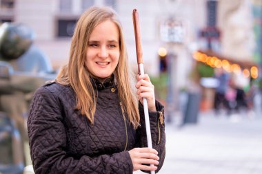 Beyaz baston tutan ve kentteki bir bankta oturan görme engelli bir kadının yakın çekimi. Gülümseyen kadın günlük kıyafetler giyiyor..  