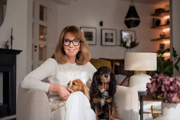 Middle-aged woman with blonde hair is sitting in an armchair at home with her cute dogs. Attractive female wearing glasses and white clothes.