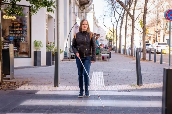 Portrait of blind woman with white cane crossing on the road in the city. A visually impaired woman wearing casual clothes and using her cane to cross the street. Full length shot.