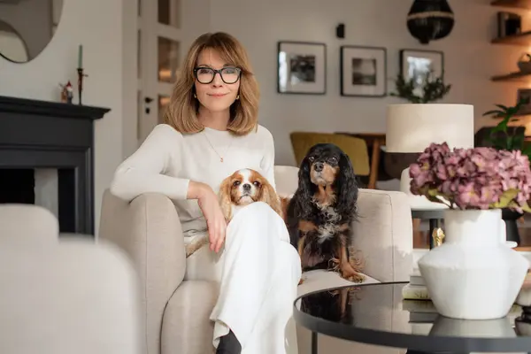 ブロンドの髪を持つ中年の女性は かわいい犬と一緒に自宅の椅子に座っています 魅力的な女性はメガネと白い服を着ています ロイヤリティフリーのストック画像
