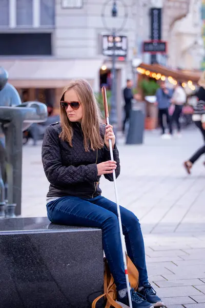 一名视力受损的妇女手持一根白色手杖坐在城市的长椅上进行了特写 穿着休闲装微笑的女人 全长射击 图库图片