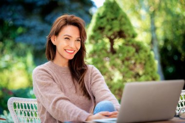 Balkonda dizüstü bilgisayarı olan kendinden emin orta yaşlı bir kadın oturuyor. Çekici kadın süveter giyiyor ve gülümsüyor.. 