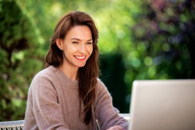 Balkonda dizüstü bilgisayarı olan kendinden emin orta yaşlı bir kadın oturuyor. Çekici kadın süveter giyiyor ve gülümsüyor.. 