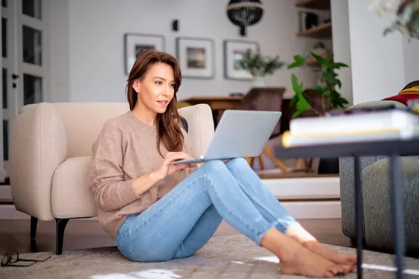 全长拍摄快乐女人使用笔记本电脑和有视频通话的同时放松在家里 穿着毛衣和蓝色牛仔裤的迷人女人 免版税图库图片