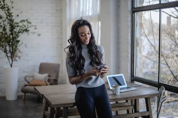 一个年轻的非洲裔美国女人站在窗前 望着别处 长发女人笑着 手里拿着智能手机 她在发短信 图库照片
