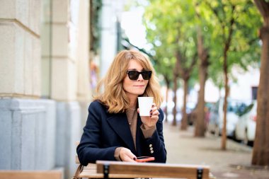 Orta yaşlı bir kadın kafenin dışında bir masada oturuyor ve bir fincan kahve içerken akıllı telefonunu kullanıyor. Gündelik kıyafetler ve güneş gözlüğü takan kendinden emin bir kadın..  