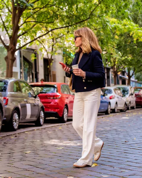 彼女の手に紙のコーヒーカップと彼女の携帯電話を持っている魅力的な中年の女性の完全な長さは 街路を歩いています 青いコートと白いズボンを身に着けている中年の女性 ストック写真