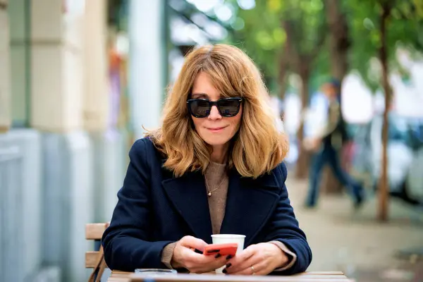 Medelålders Kvinna Som Sitter Utanför Caféet Och Använder Sin Smartphone Stockbild