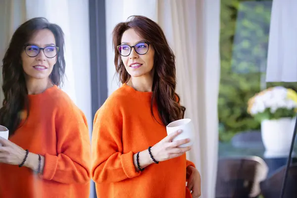 窓に立っている茶色の髪の女性が手元にコーヒーカップを置いています オレンジ色のセーターを着て窓の外を見ている中年の女性 ストックフォト