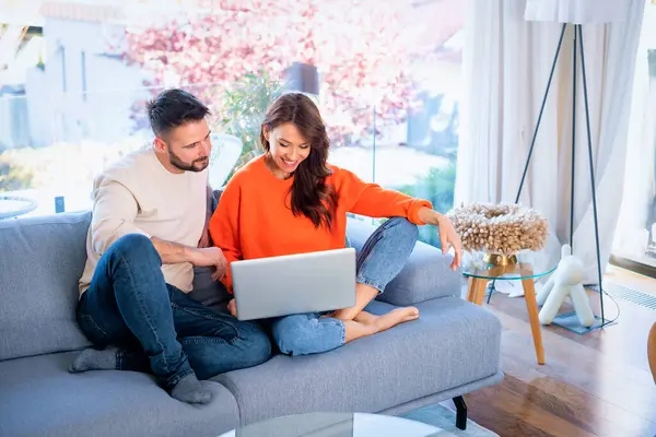 女性と男性は 自宅でソファーに座っている間 ノートパソコンを使用しています ウェブを閲覧する幸せなカップル カジュアルな服を着ている ストック画像