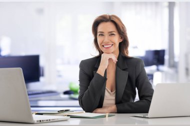 Esmer, orta yaşlı iş kadını ofiste oturuyor ve iş için dizüstü bilgisayar kullanıyor. İş kıyafeti giyen kendine güvenen profesyonel bir kadın..