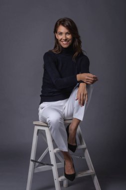 Esmer, orta yaşlı bir kadının soyutlanmış gri arka planda oturması. Siyah balıkçı yaka kazak giyen ve neşeli gülümseyen çekici bir kadın. Boşluğu kopyala.