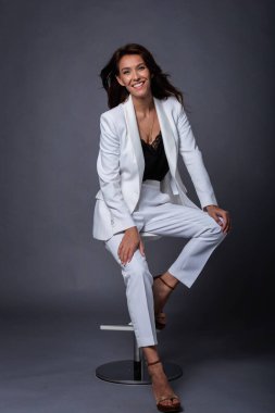 Esmer, orta yaşlı bir kadının soyutlanmış gri arka planda oturması. Beyaz takım elbiseli çekici bir kadın ve neşeli bir gülümseme. Boşluğu kopyala. 