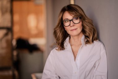 Duvara yaslanmış orta yaşlı sarışın bir kadının portresi. Beyaz gömlek ve gözlük takan çekici bir kadın.. 