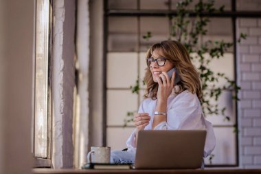 Elinde kupa olan orta yaşlı sarışın bir kadın düşünceli bir şekilde arama yapıyor. Beyaz gömlek giyen ve gülümseyen çekici bir kadın. Merkez Ofis. 