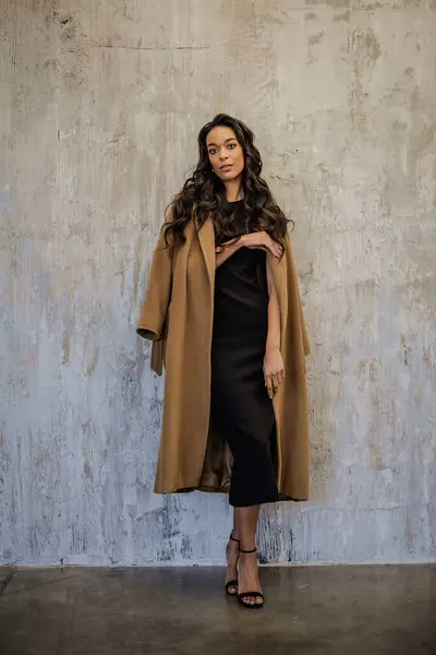 stock image An African American woman standing in front of a grey wall. She is looking the camera and wearing a black dress and camel coat. Copy space. Full length.