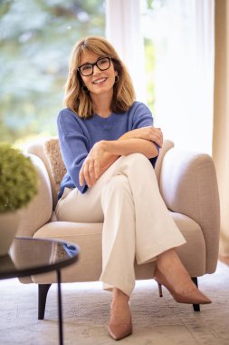 Sarışın, orta yaşlı bir kadının boyu evde bir koltukta oturuyor. Mavi gömlekli, beyaz pantolonlu ve gülümseyen kendinden emin bir kadın.. 