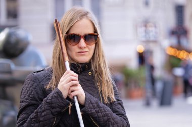 Beyaz baston tutan ve sokakta duran görme engelli bir kadının yakın çekimi. Gülümseyen kadın günlük kıyafetler giyiyor.. 