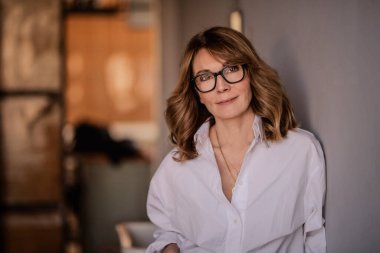 Duvara yaslanmış orta yaşlı sarışın bir kadının portresi. Beyaz gömlek ve gözlük takan çekici bir kadın.. 