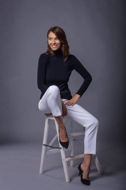 Gri bir duvarın önünde oturan çekici kahverengi saçlı kadın. Siyah balıkçı yaka kazak giyen ve gülümseyen orta yaşlı bir kadın. Stüdyo çekimi. İzole edilmiş arka plan. Tam boy çekim.