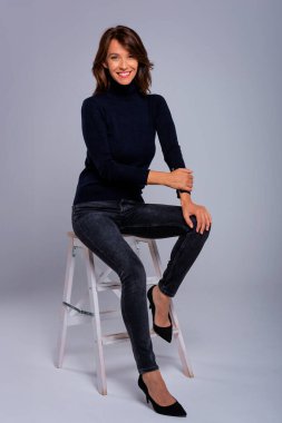 Gri bir duvarın önünde oturan çekici kahverengi saçlı kadın. Siyah balıkçı yaka kazak giyen ve kameraya gülümseyen orta yaşlı bir kadın. Stüdyo çekimi. İzole edilmiş arka plan. Tam boy çekim.