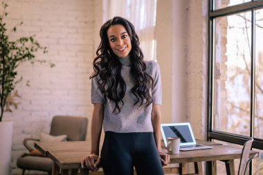 Masada durmuş kameraya bakan Afro-Amerikalı bir kadın. Rahat kıyafetler giyen kendine güvenen bir kadın.. 