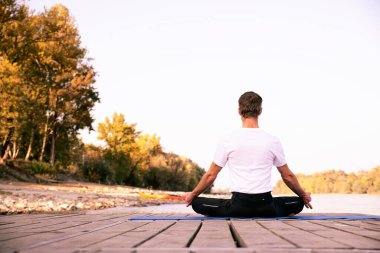 Dışarıda yoga yapan orta yaşlı bir adam. Kafkas adam yoga minderi kullanıyor ve doğa üzerinde nilüfer pozu veriyor. Arka plan görüntüsü.