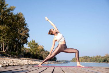 Full length of caucasian woman practicing yoga on pier by the river. Attractive female has blond hair and smiling.  clipart