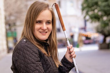 Beyaz baston tutan ve sokakta duran görme engelli bir kadının yakın çekimi. Gülümseyen kadın günlük kıyafetler giyiyor.. 