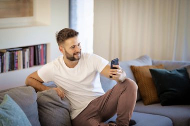 Orta yaşlı bir adam evdeki kanepede oturuyor ve telefon kullanıyor. Sıradan erkek mesajlaşması.