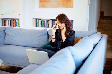 Orta yaşlı bir kadın evinde kanepede oturuyor ve dizüstü bilgisayarını ve cep telefonunu kullanıyor. Evde çalışan kendine güvenen bir kadın. Merkez Ofis