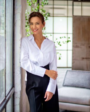 Pencerenin önünde duran ve kameraya gülümseyen orta yaşlı güzel bir kadının portresi. Esmer kadın, beyaz gömlek ve siyah pantolon giyiyor..