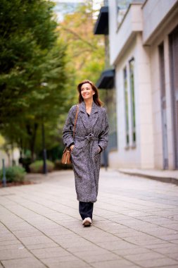 Sonbahar günü şehir caddesinde yürüyen esmer saçlı orta yaşlı bir kadın. Tüvit ceket giyiyor..