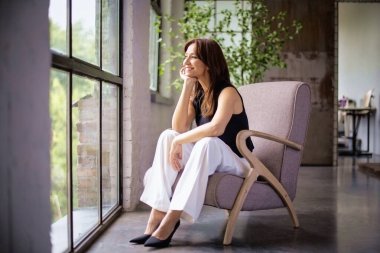 Çekici, orta yaşlı kadın pencerenin kenarında dinleniyor. Kumral kadın, koltukta oturuyor ve siyah gömlek ve beyaz pantolon giyiyor..