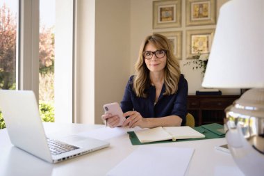 Masada oturmuş, dizüstü bilgisayar ve akıllı telefon kullanan gülümseyen olgun bir kadın. Evde çalışan sarı saçlı bir kadın.. 
