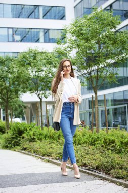 Şehirde yürürken akıllı telefonunu kullanan gülümseyen bir kadın. Esmer saçlı iş kadını, telefonda biriyle konuşuyor. Tam uzunluk. 